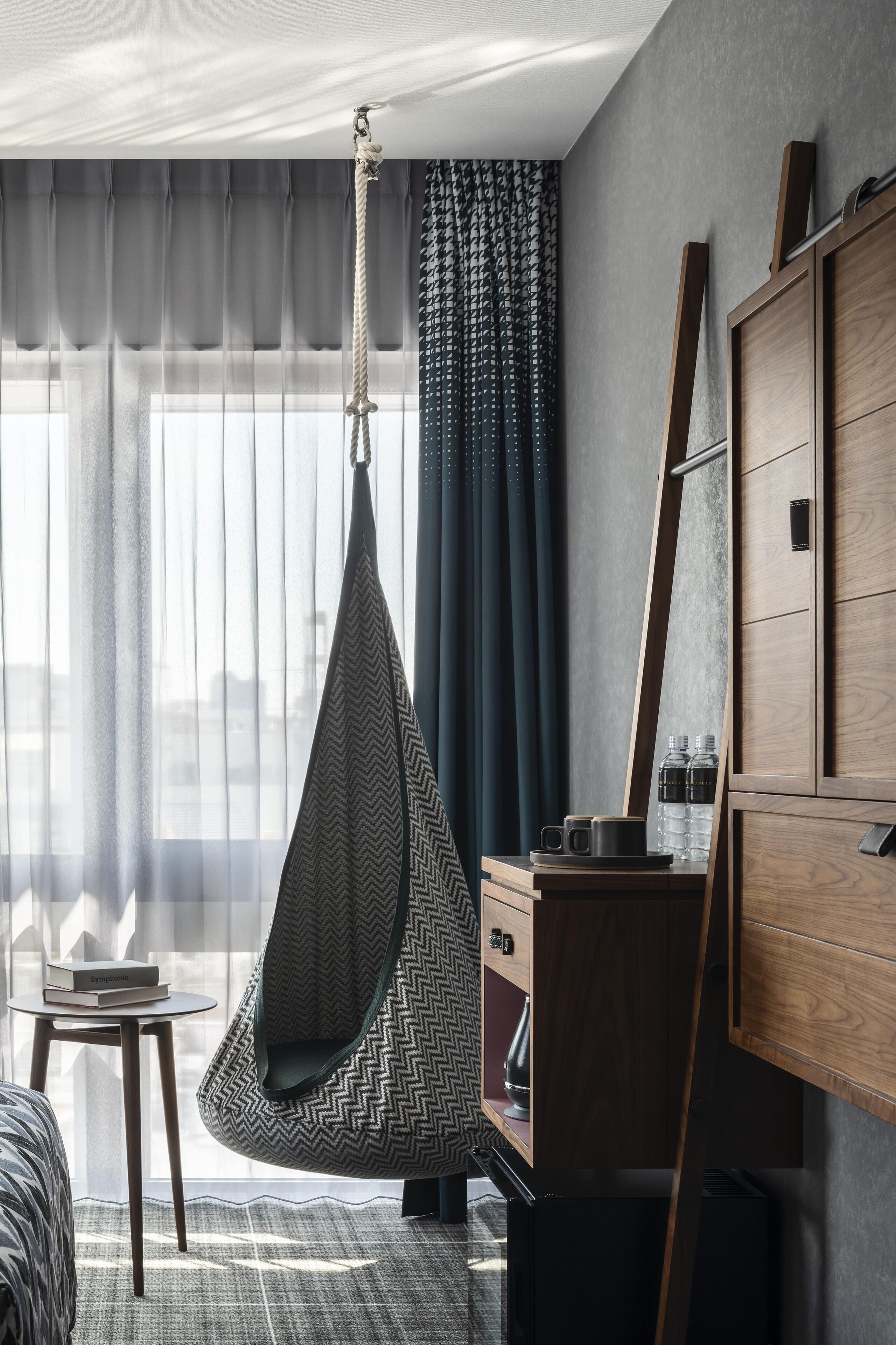 Een moderne hotelkamer met een hangstoel, een klein tafeltje met boeken, een kledingkast en uitzicht op de stad door dunne gordijnen.