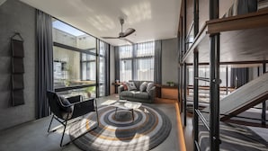 Uma sala de estar moderna com um sofá, uma mesa de centro e uma poltrona preta. Há grandes janelas e um ventilador de teto.
