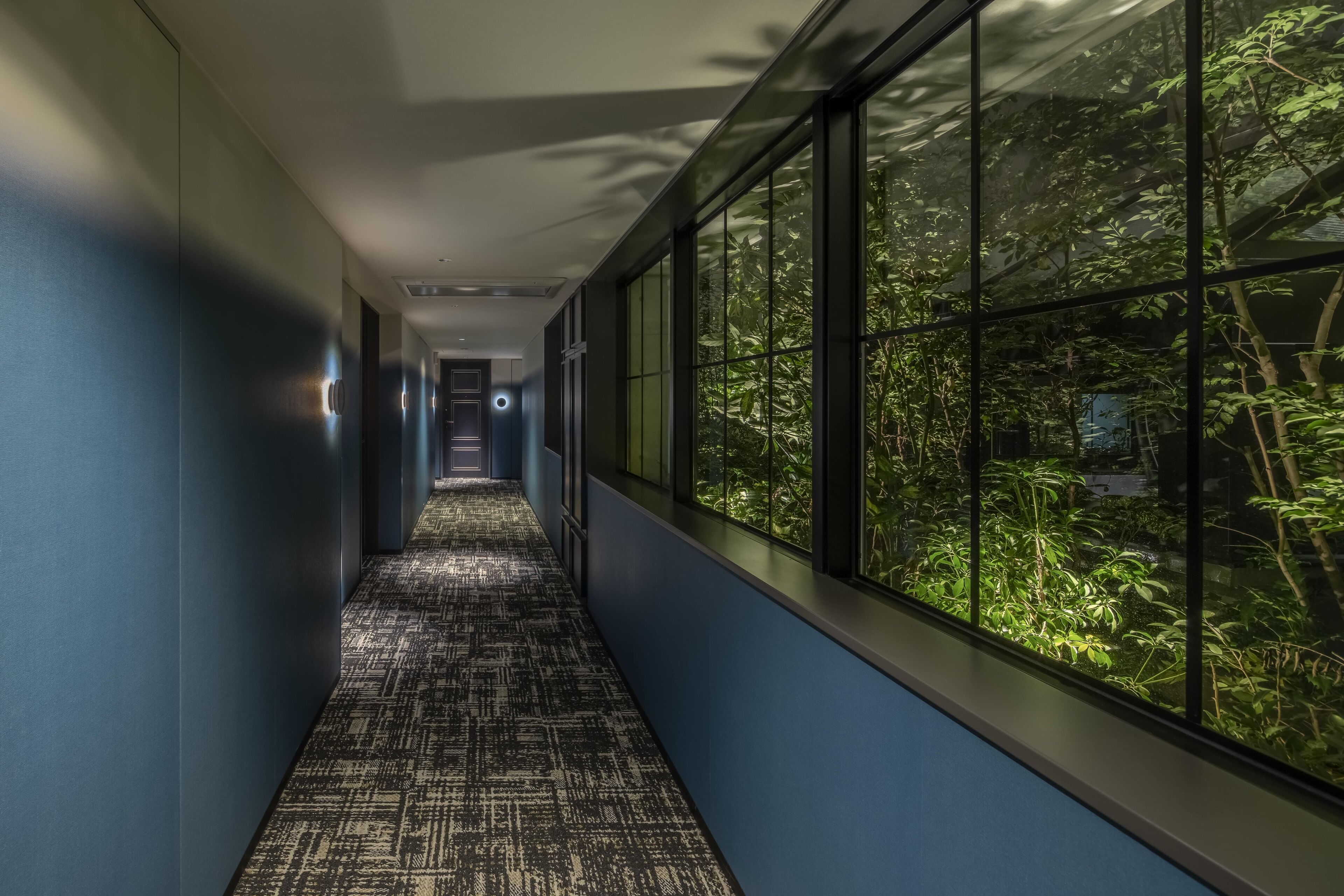 A hallway with patterned carpet, blue walls, and large windows overlooking greenery.