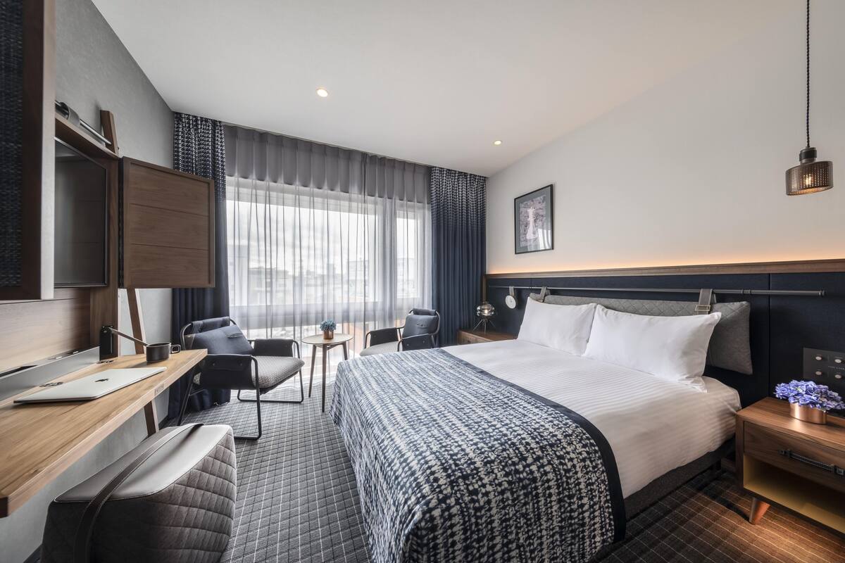 A modern hotel room with a large bed, a desk with a laptop, a chair, a small table with a plant, and a nightstand with a lamp.