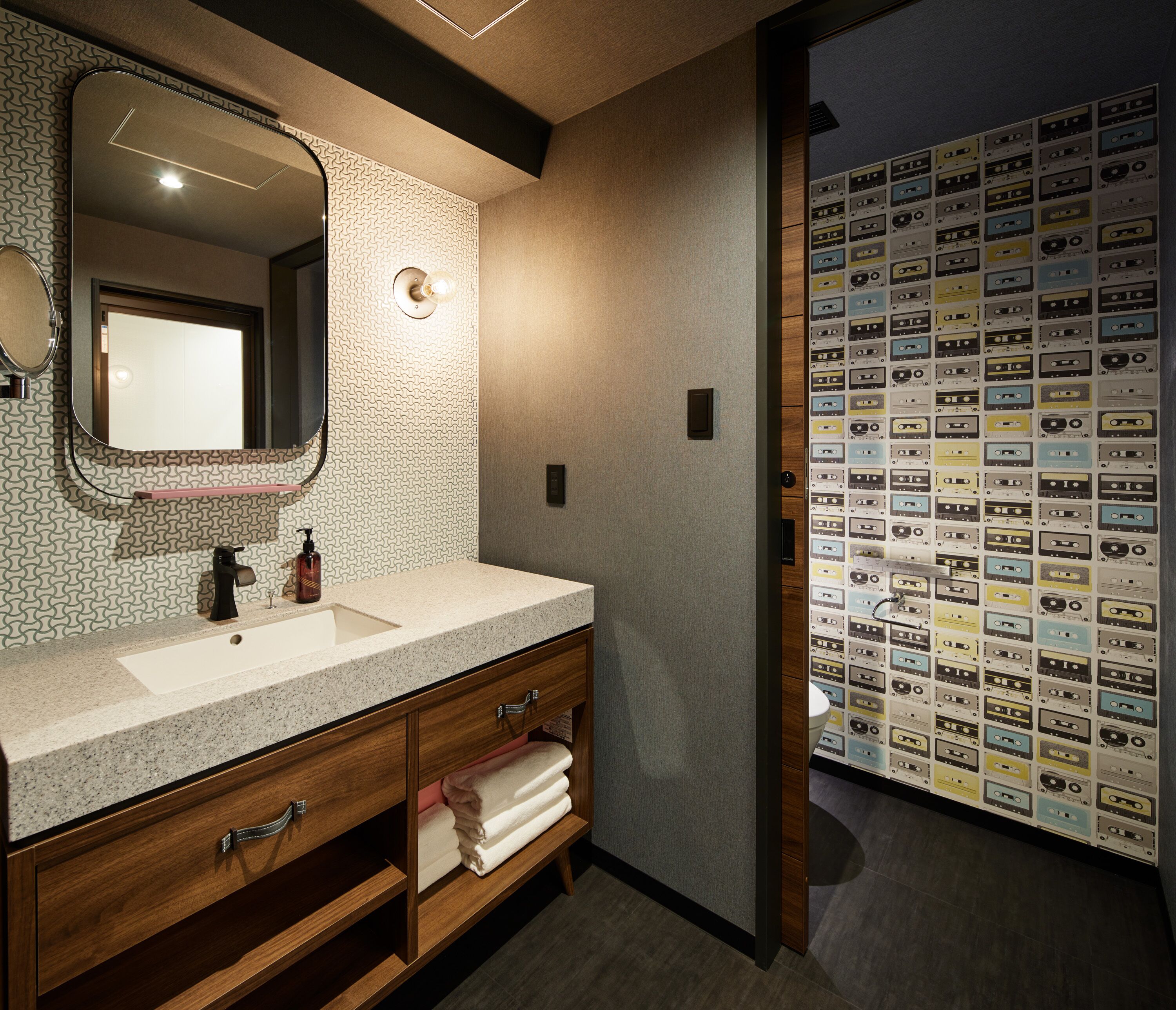 A modern bathroom with a granite countertop, wooden cabinetry, a large mirror, and a wall of vintage cassette tapes.