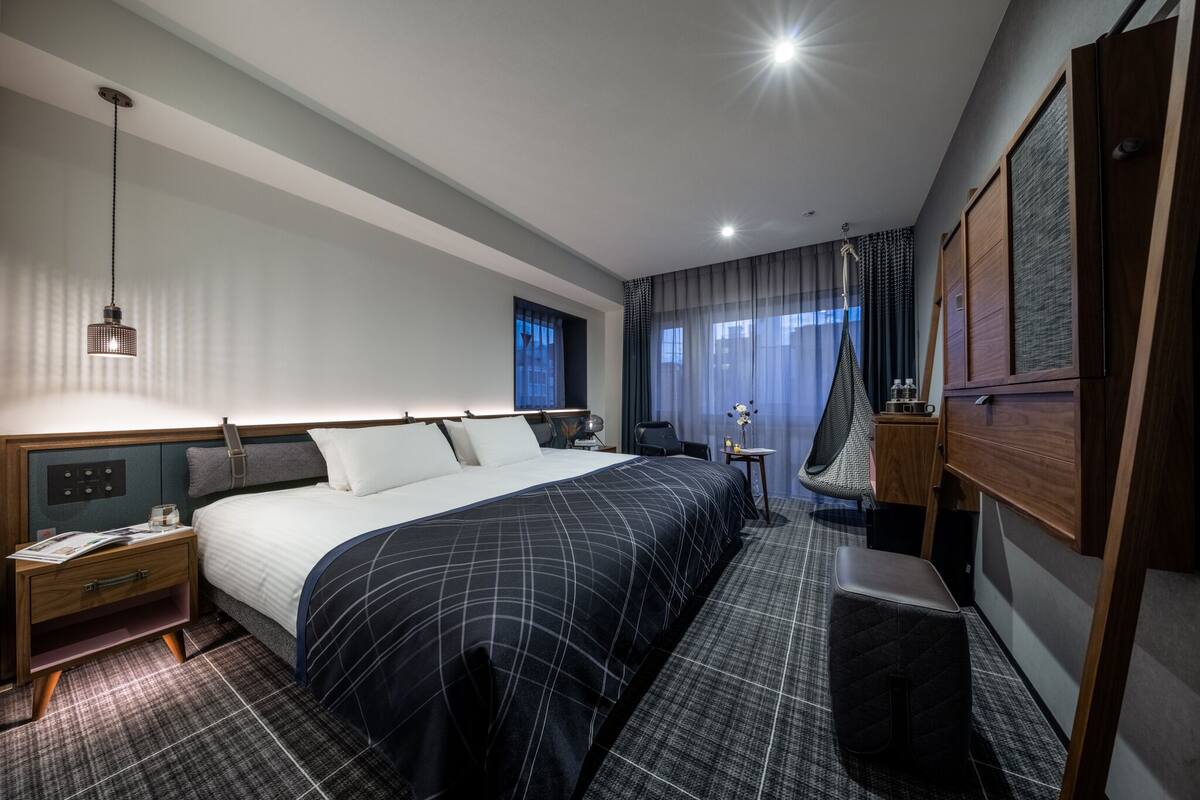A modern hotel room with a large bed, a bedside table, a desk, a chair, a fan, and a window with curtains.
