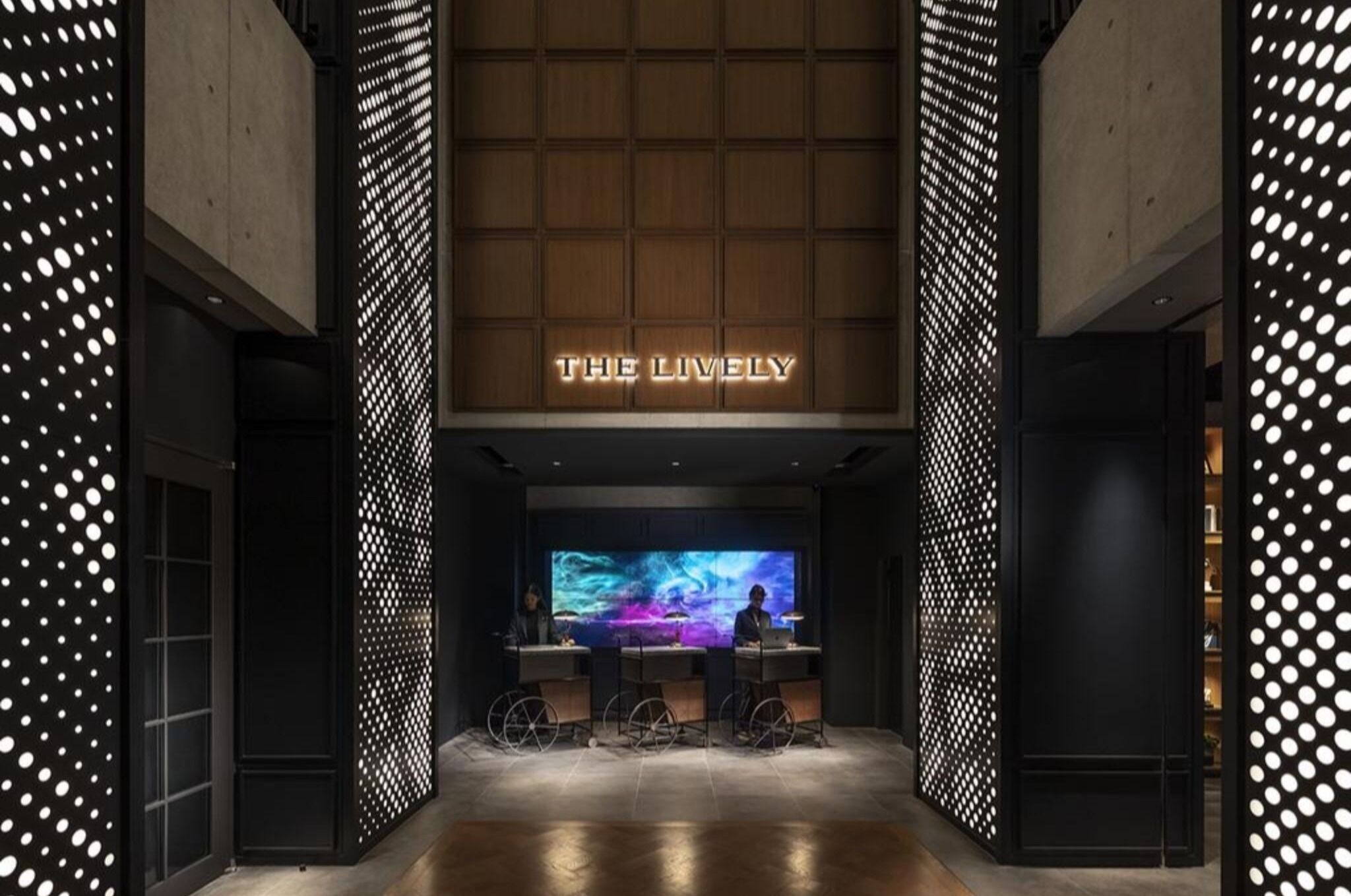 A modern hotel lobby with a large digital screen, a reception desk, and a sign that reads 'THE LIVELY'.