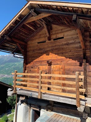 Una cabaña de madera con balcón, barandilla metálica y vistas a un paisaje montañoso.