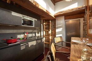 A compact kitchen with modern appliances, a dining area with a wooden table and chairs, and a stone accent wall. - ATTIC VALAISAN TYPICAL (Mase)