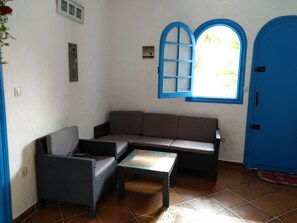 Ein Zimmer mit einem braunen Sofa, zwei Korbstühlen, einem kleinen Glastisch, einem Fenster mit blauen Rahmen und einer blauen Tür.