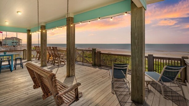 A wooden deck with a view of the ocean, featuring a swing, chairs, and a table.