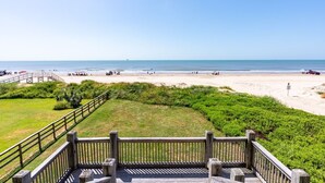 A beach view from a wooden deck with a railing, leading to a sandy beach area with a wooden walkway and some greenery.
