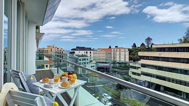 A balcony with a table set for breakfast, overlooking a cityscape with modern buildings and a clear sky. - Apartment Sunniva by LovelyStay (Funchal)