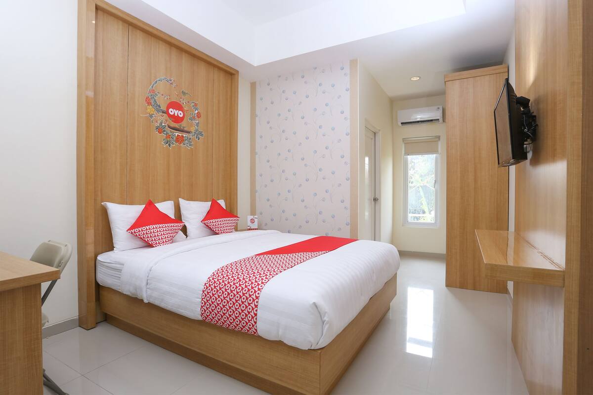 Habitación de hotel con cama de madera, ropa de cama blanca, almohadas rojas y blancas, televisor montado y armario de madera.