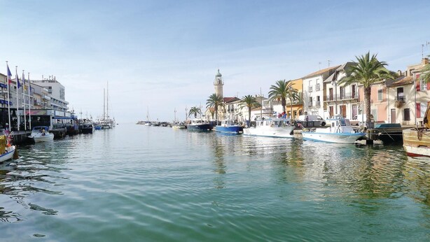 Un port de plaisance avec des bateaux amarrés le long d’une promenade, des bâtiments dotés de balcons et une tour imposante en arrière-plan.