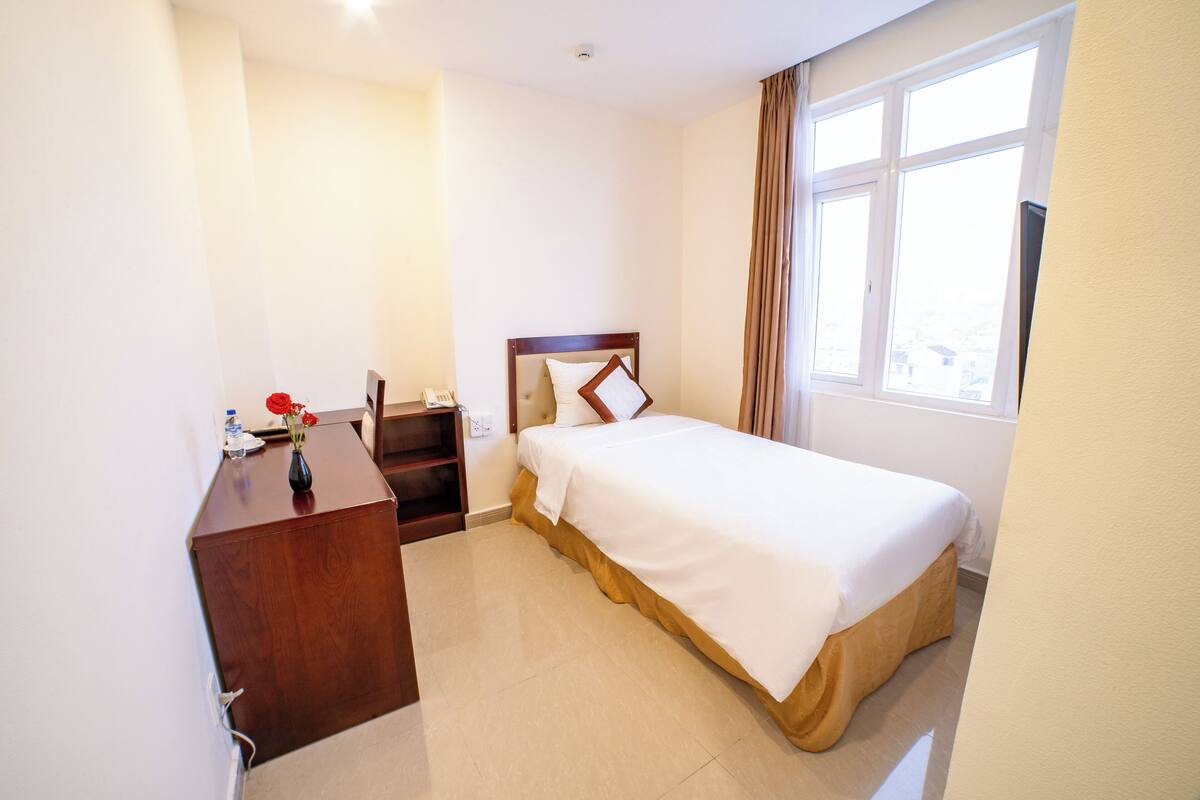 A hotel room with a bed, a wooden desk, a vase with flowers, and a window with curtains.