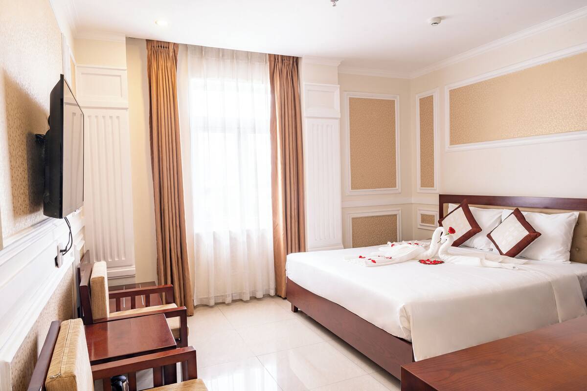 A hotel room with a large bed, a flat-screen TV mounted on the wall, a wooden desk, and a window with curtains.