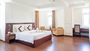 A spacious hotel room with a large bed, a sitting area with a chair and table, and a desk with a chair. - Lychee Hotel (Da Nang)