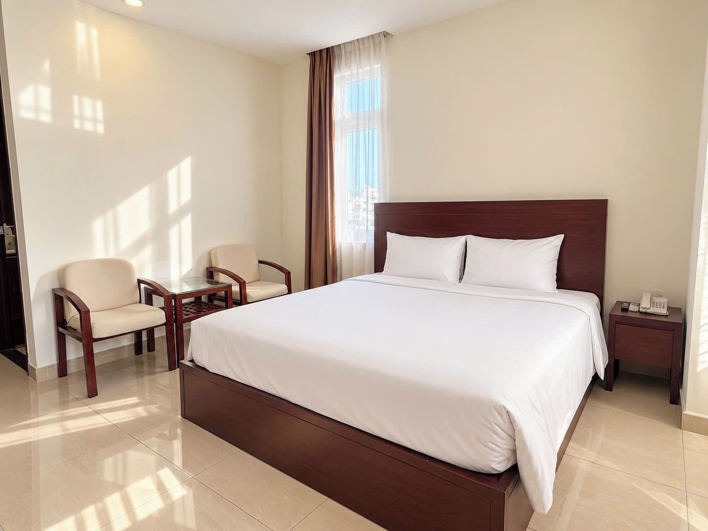 A hotel room with a large bed, two armchairs, a small table, a nightstand with a phone, and a window with curtains.