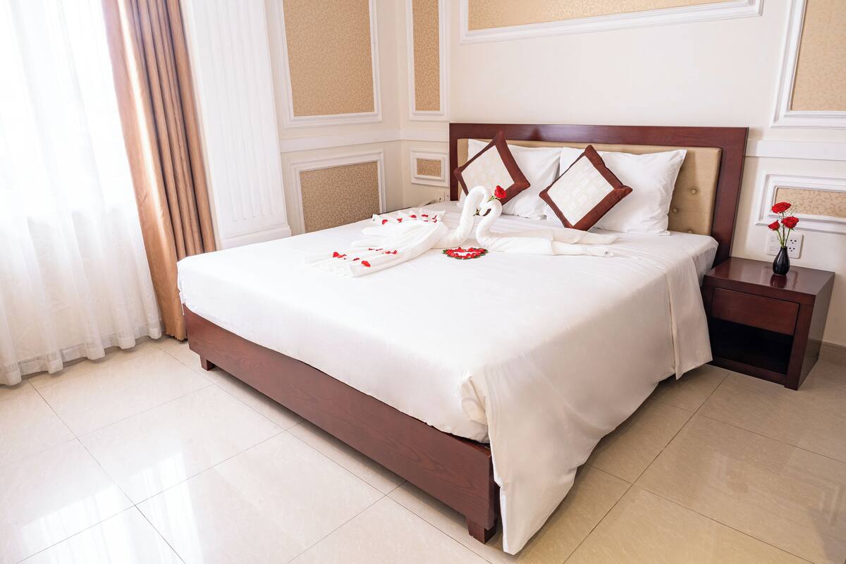 A neatly made bed with white linens and decorative swan-shaped pillows, a wooden nightstand with a vase of red flowers, and a light-colored tiled floor.
