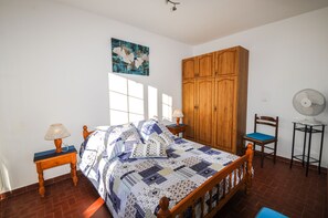 A bedroom with a wooden bed, a nightstand with a lamp, a wooden wardrobe, a chair, and a standing fan.