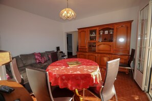 A dining area with a round table covered in a red tablecloth, surrounded by chairs. A wooden cabinet with glass doors is visible in the background.