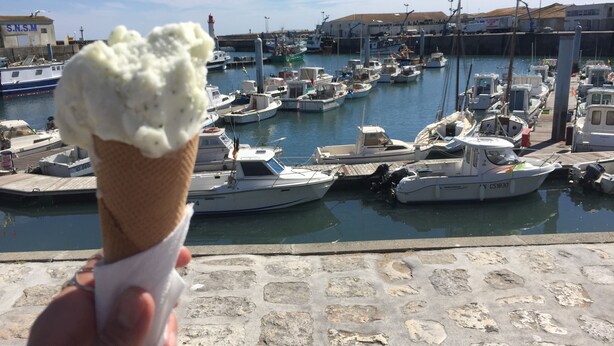 Una mano sosteniendo un cono de helado, con un puerto deportivo al fondo.