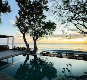 An infinity pool reflecting the sky and trees, with a wooden deck and lounge chairs.