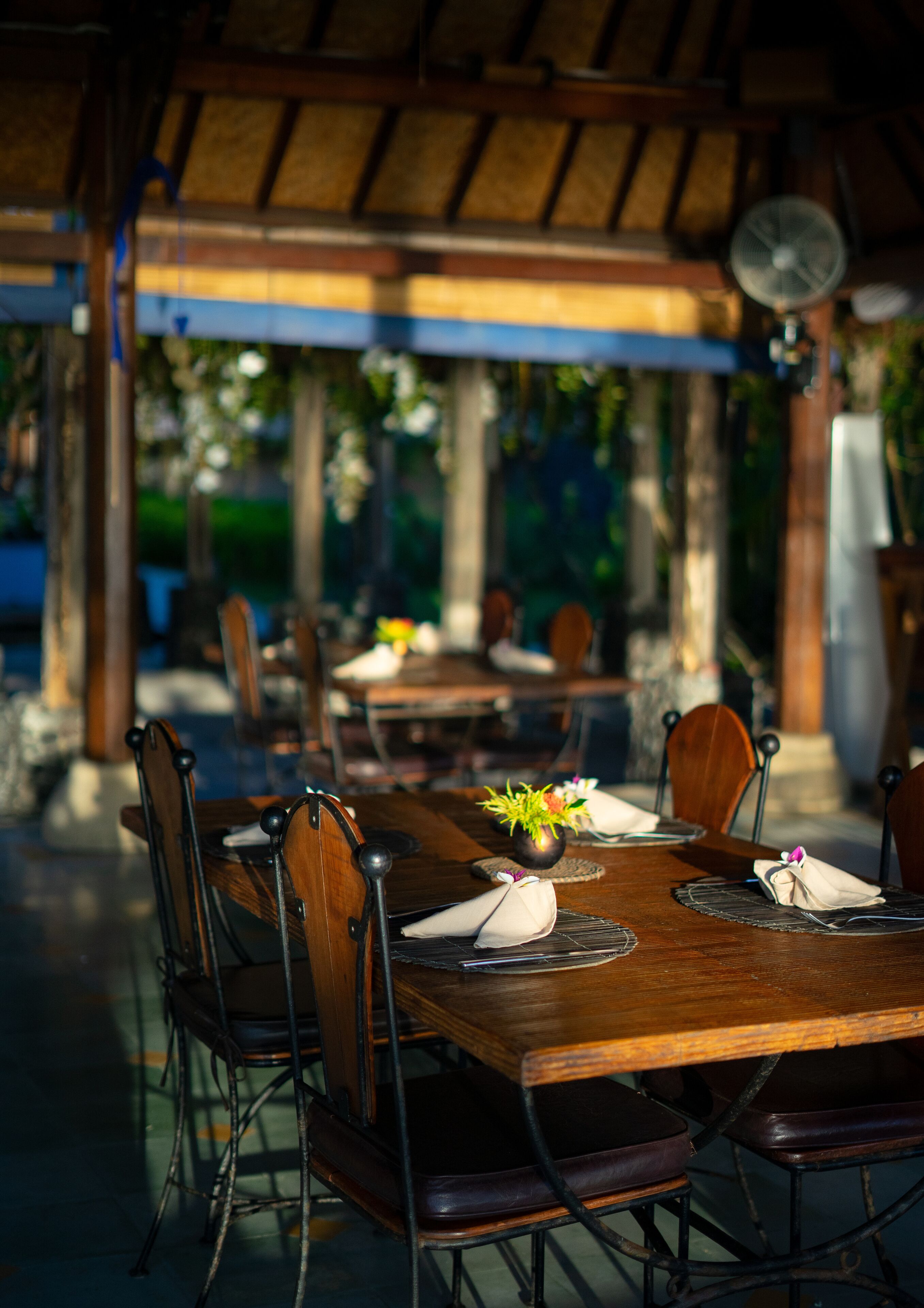 Un área de comedor al aire libre con mesas y sillas de madera, techo de paja y un ventilador.