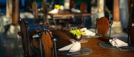 An outdoor dining area with wooden tables and chairs, a thatched roof, and a fan.