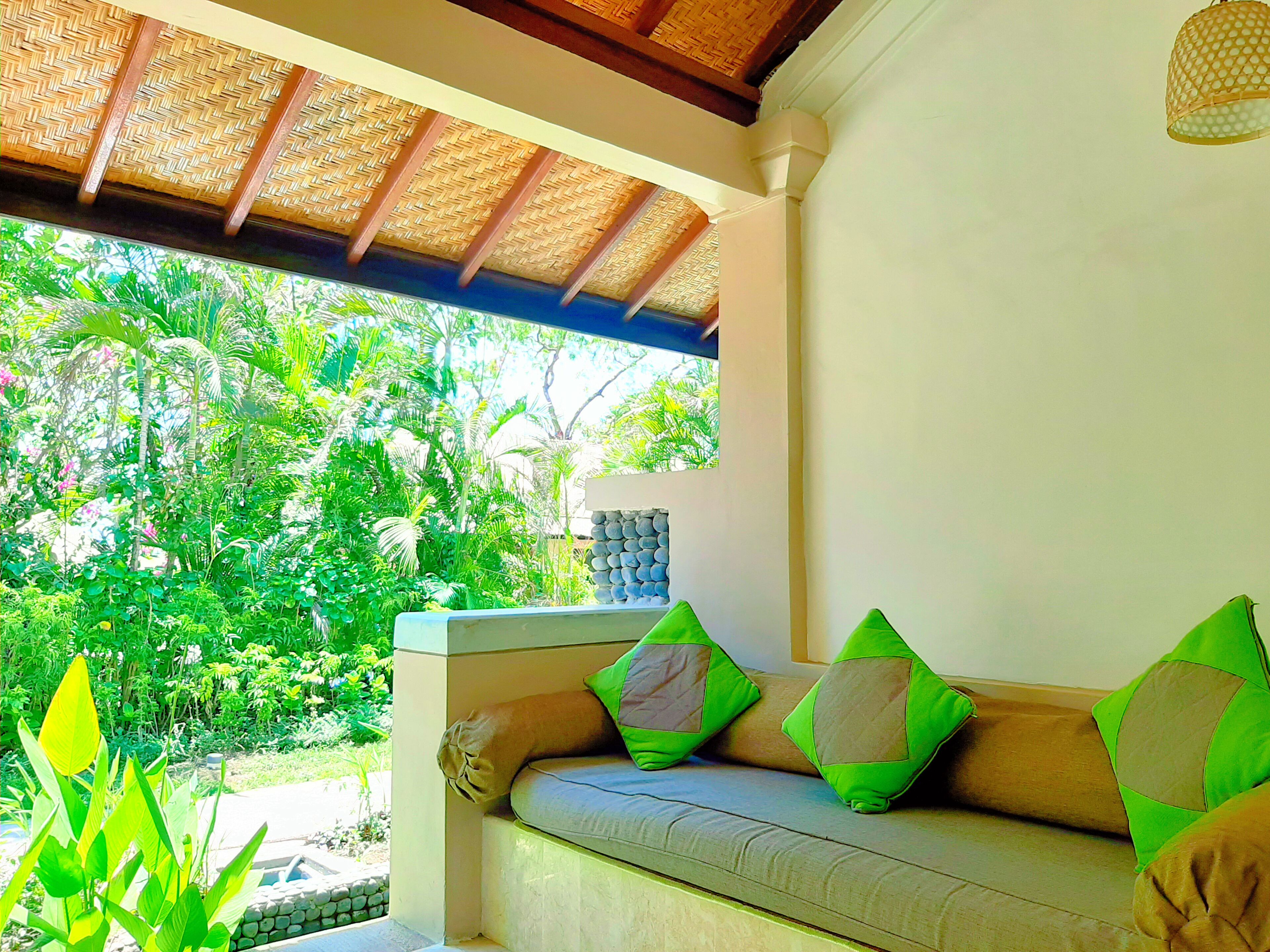 A room with a sofa, green cushions, a woven ceiling, and a view of lush greenery outside.
