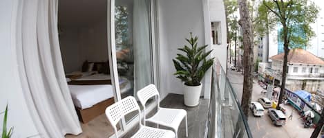 Un balcón con una cama, dos sillas blancas, una planta en maceta y vista de la calle.