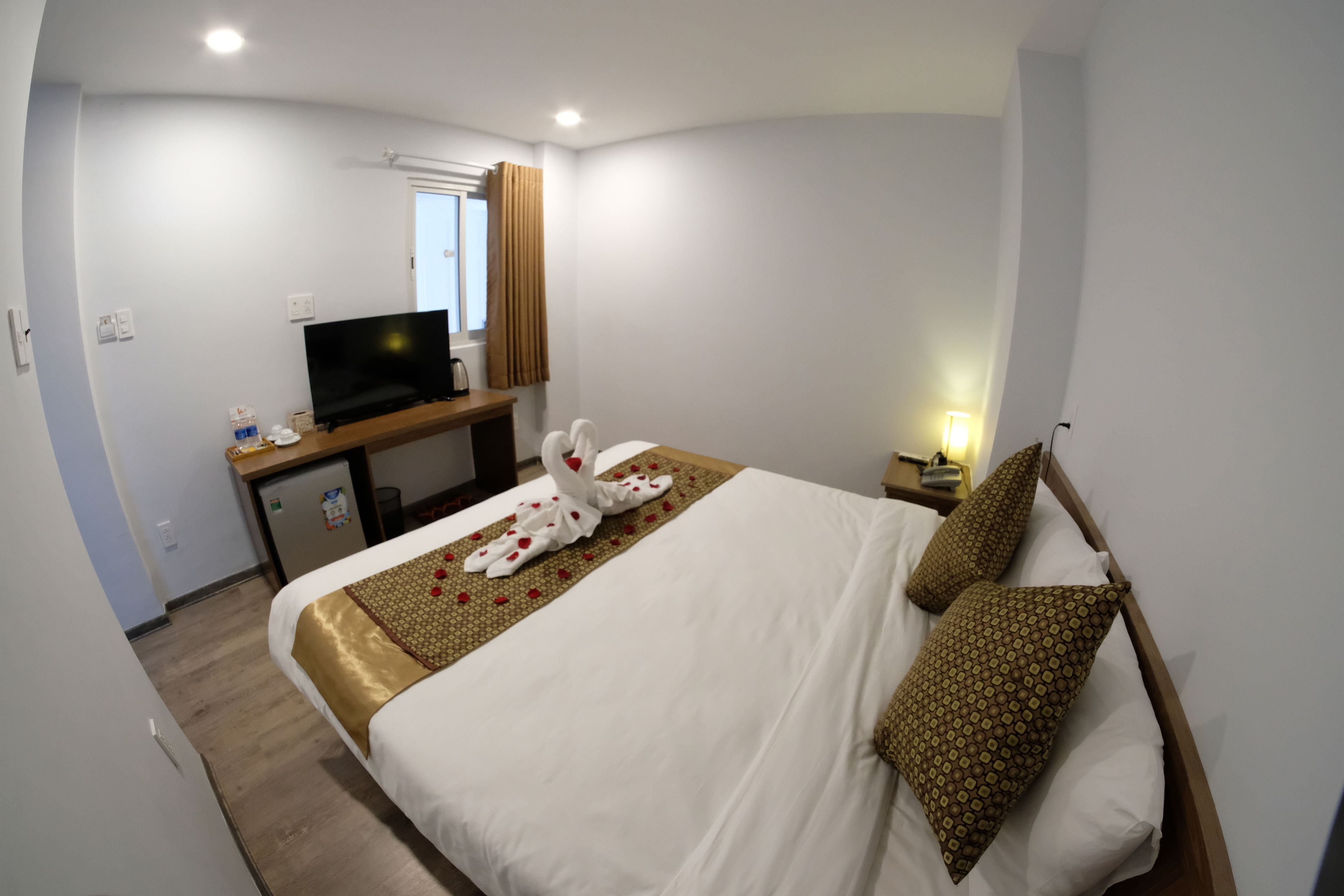 A hotel room with a bed, a desk with a television, a small refrigerator, and a window with curtains.