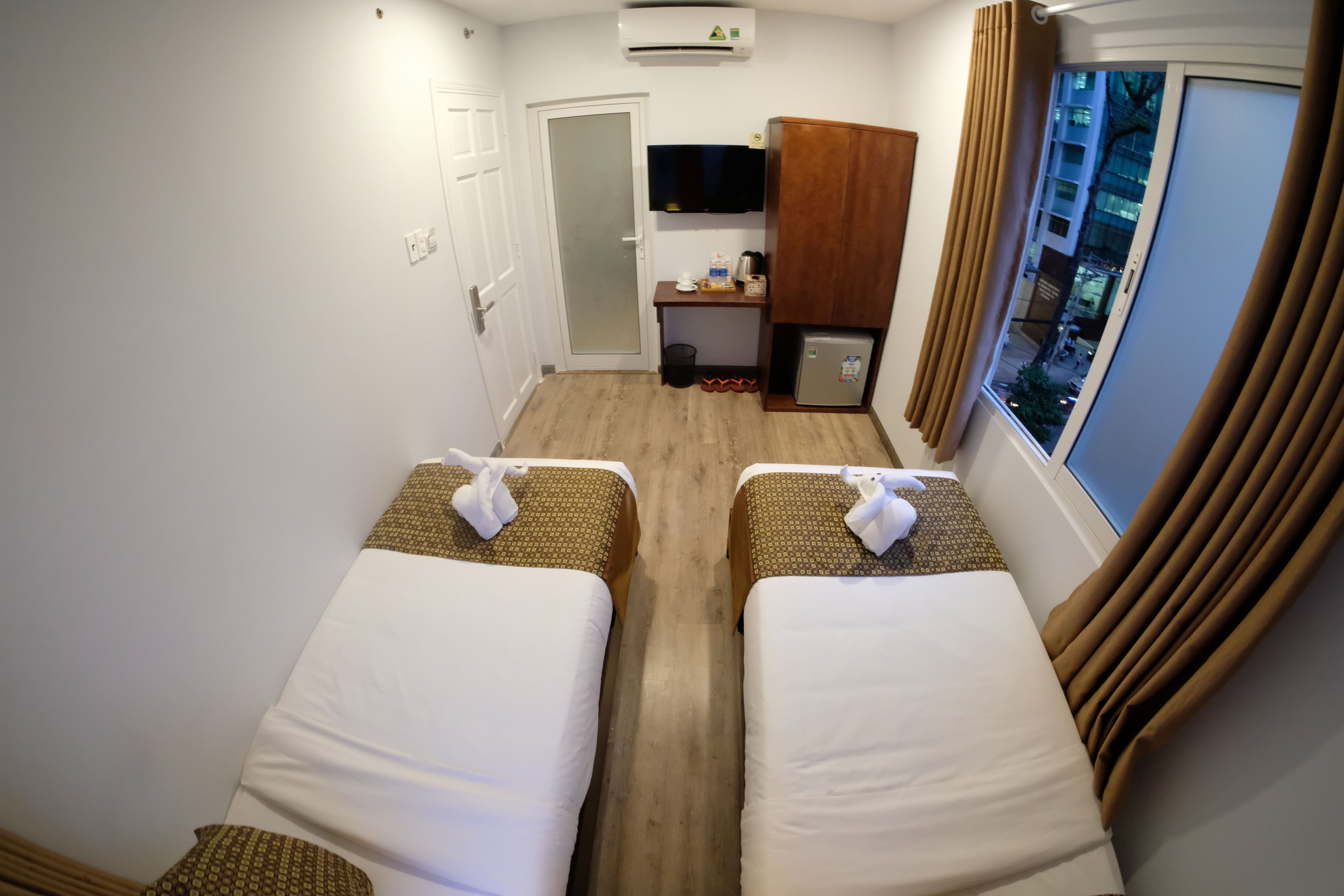 A hotel room with two beds, a small kitchenette, a TV, and a window with curtains.