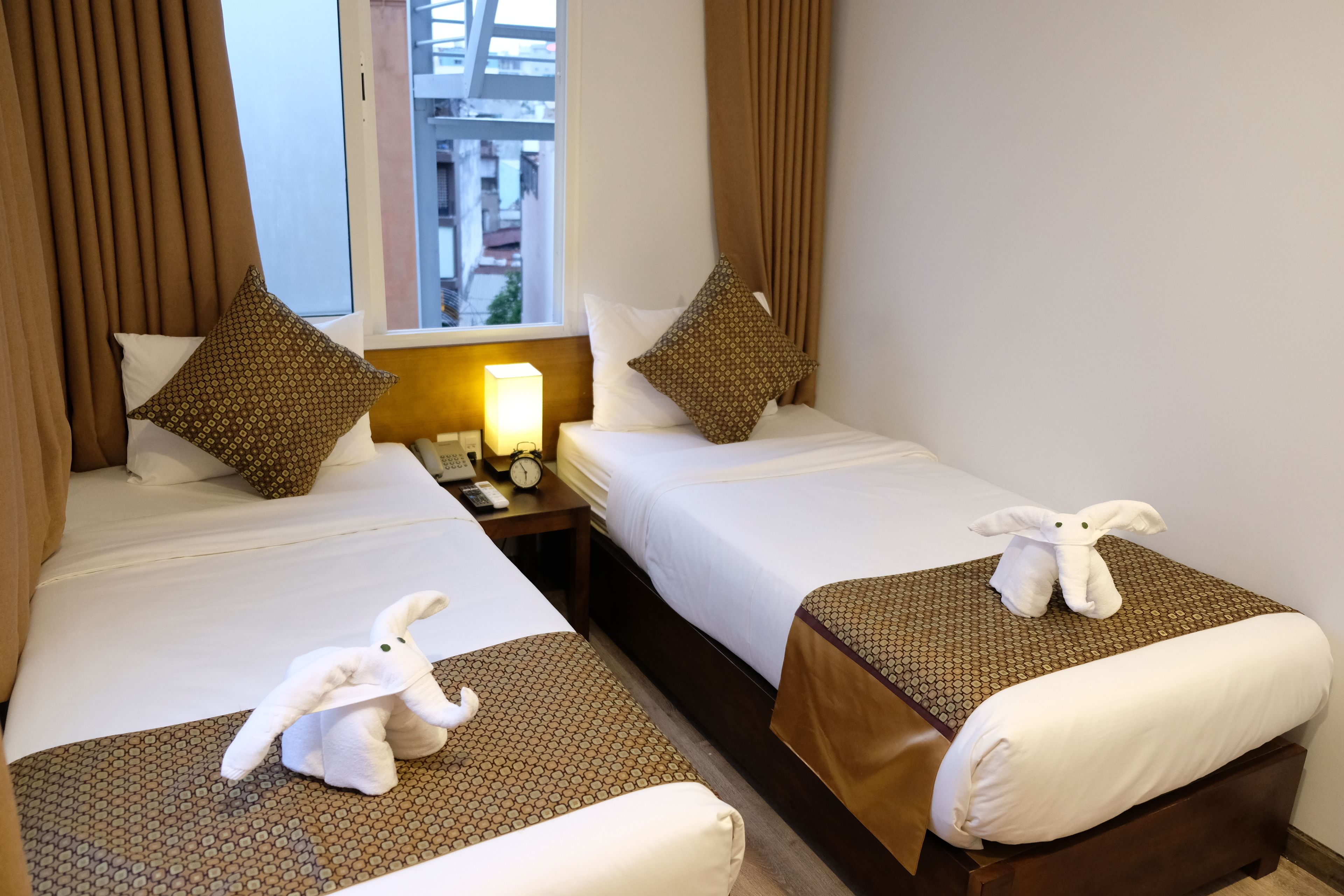 Dos camas de hotel juntas con cubrecamas estampados, almohadas y una mesita de noche con lámpara y reloj.