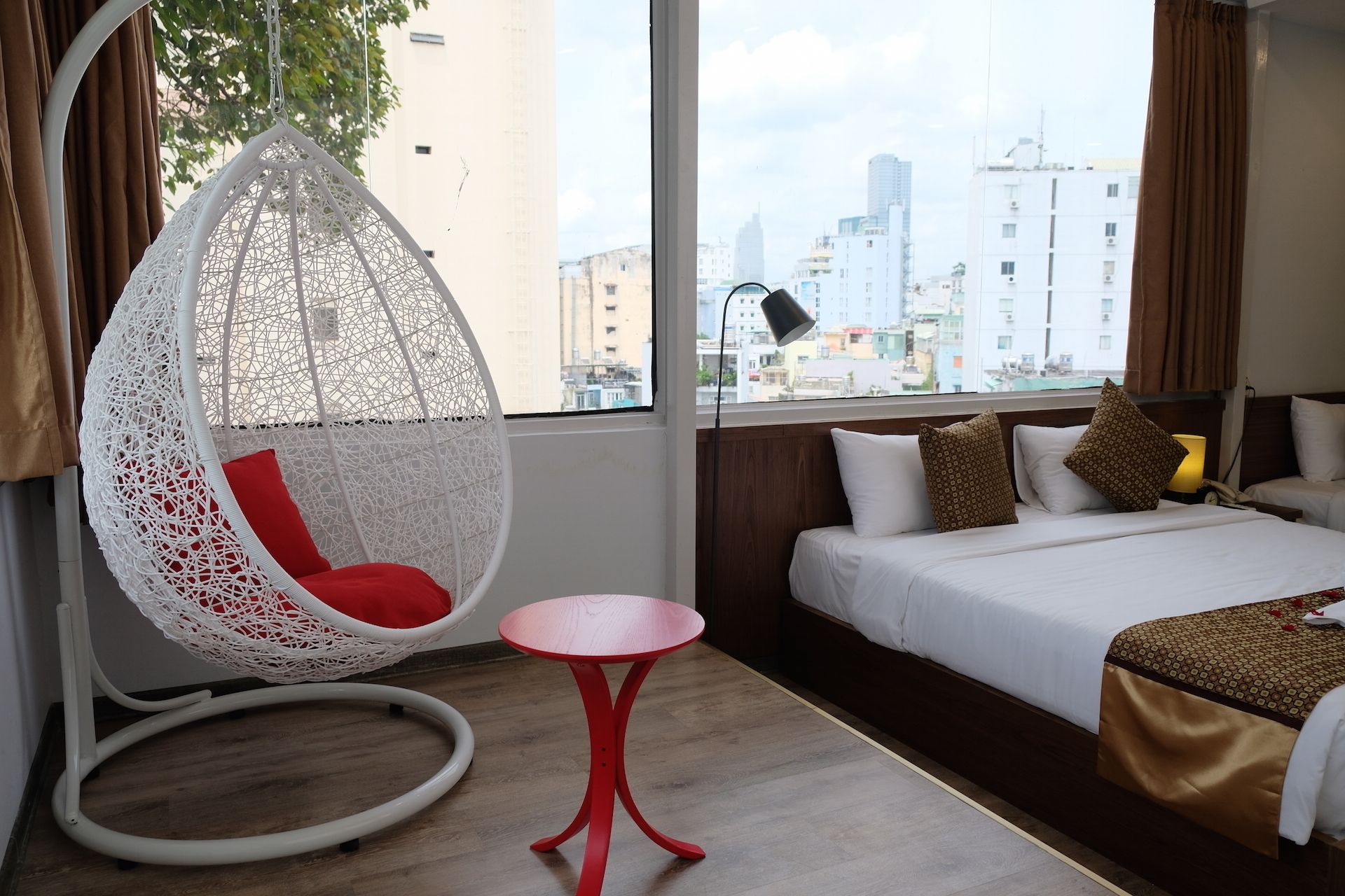 Ein Hotelzimmer mit großem Fenster, einem Hängesessel, einem kleinen Tisch, einem Bett mit gemusterten Kissen und Blick auf die Stadt.