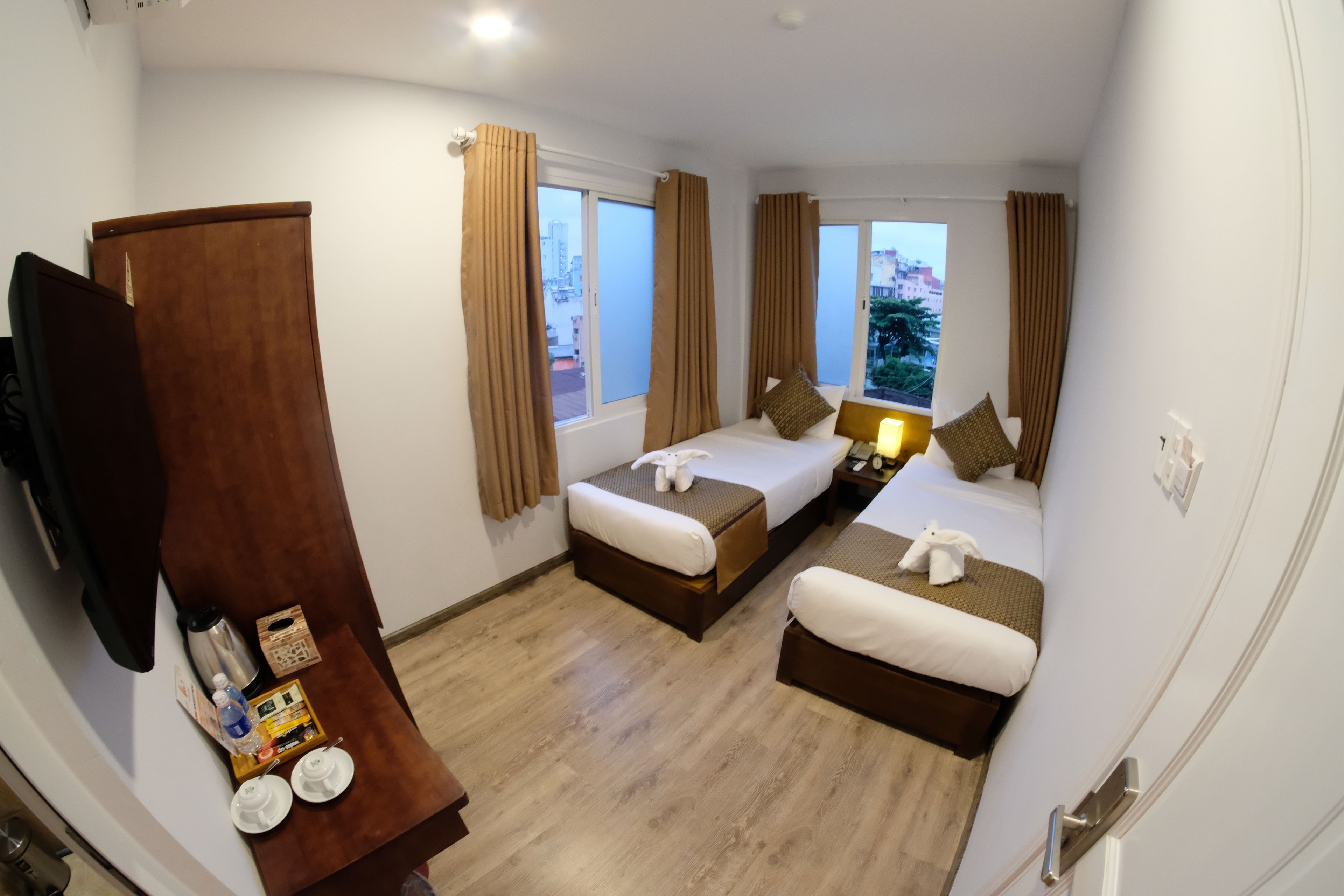 A hotel room with two single beds, a wooden wardrobe, a small table with a lamp, and a view of the cityscape through the windows.