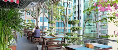 An outdoor dining area with wooden tables and chairs, surrounded by potted plants and trees.
