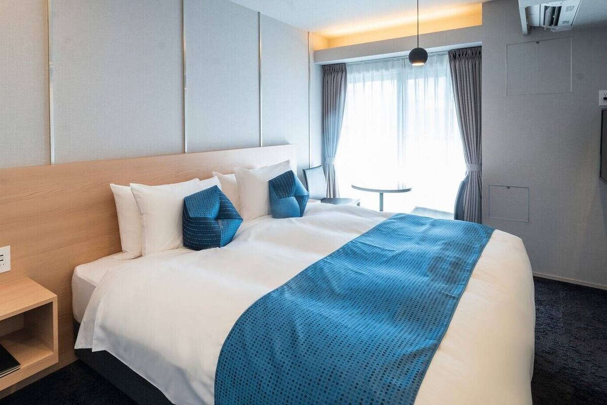 A hotel room with a bed, bedside table, wall-mounted TV, and a window with curtains.