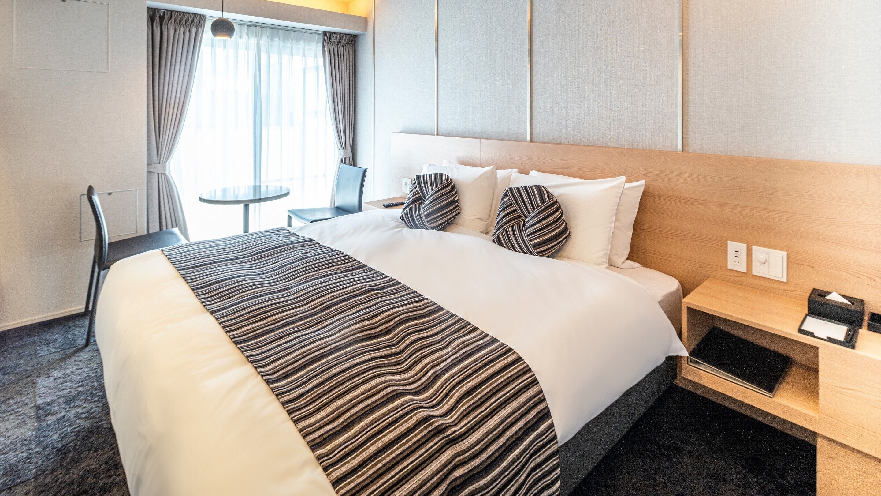 Hotellihuone, jossa on sänky, yöpöytä, tuoli, pieni pöytä ja ikkuna, jossa on verhot.