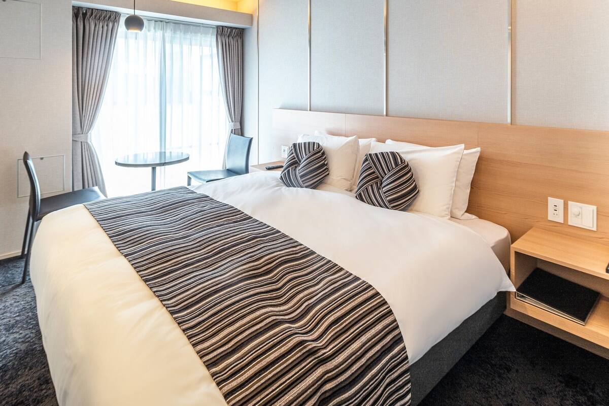 A hotel room with a bed, a bedside table, a chair, a small table, and a window with curtains.