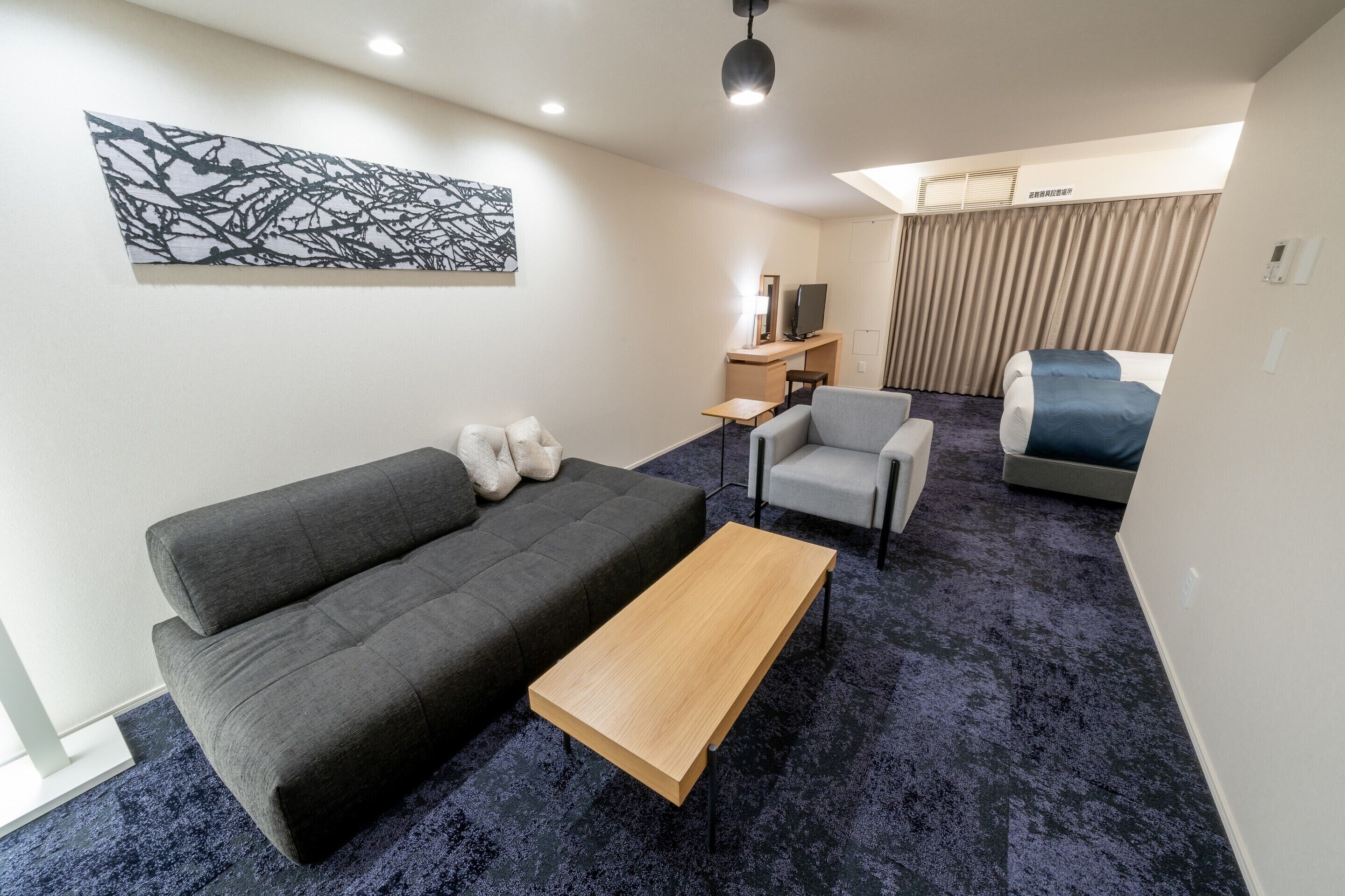 A modern hotel room with a grey sofa, a wooden coffee table, a small desk with a TV, a chair, and a bed with a blue cover.