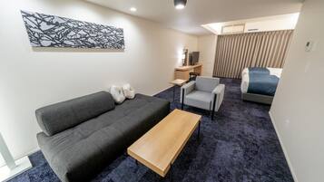 A modern hotel room with a grey sofa, a wooden coffee table, a small desk with a TV, a chair, and a bed with a blue cover.