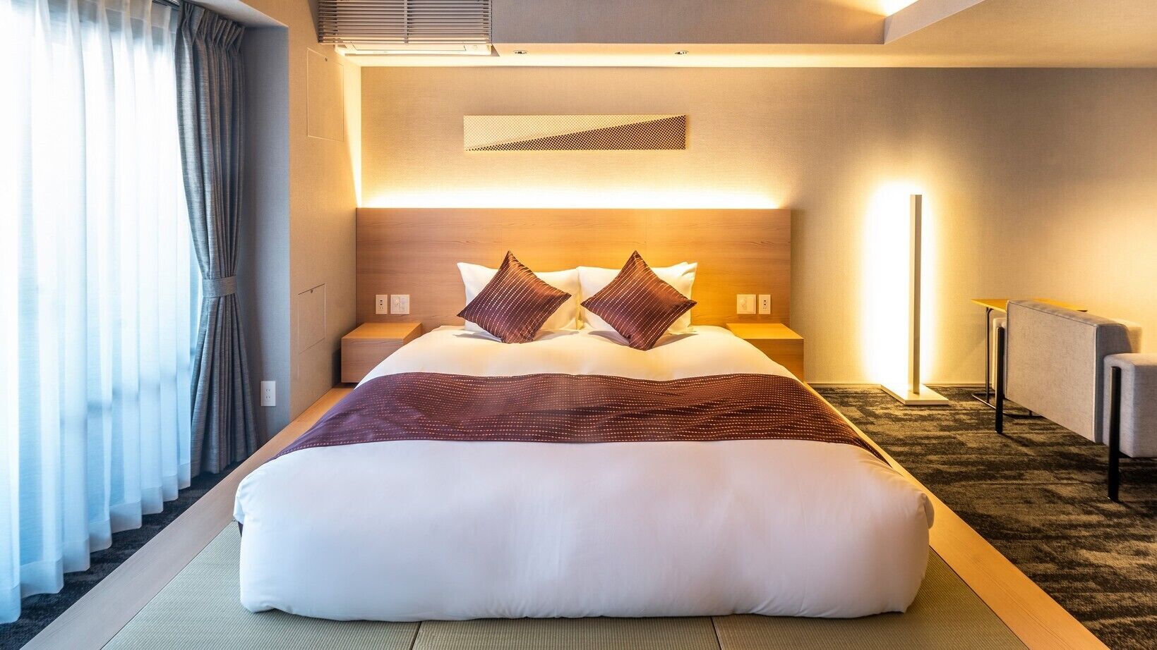 Hotellihuone, jossa on suuri sänky, yöpöydät, sängynpääty, jossa on sisäänrakennettu valaistus, näkymä ulos läpikuultavien verhojen läpi sekä matolla peitetty lattia.