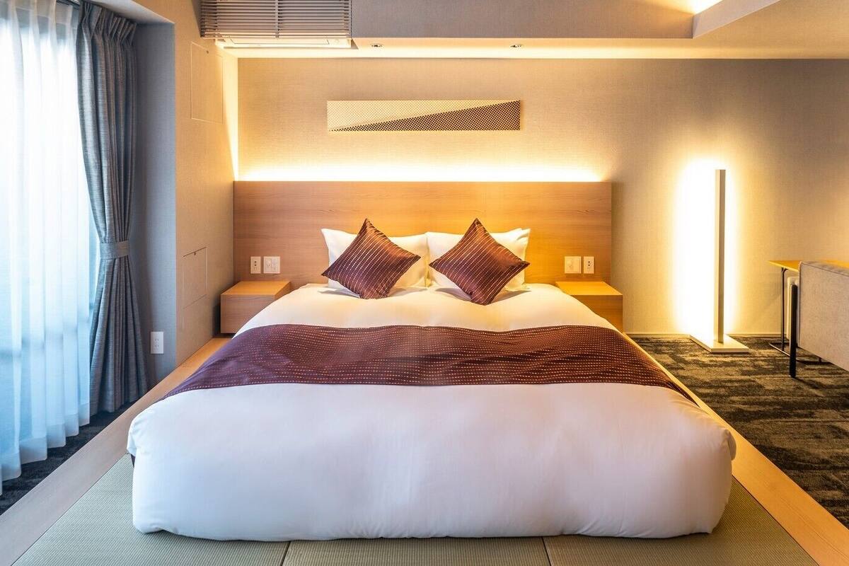 A hotel room with a large bed, bedside tables, a headboard with built-in lighting, a view of the outside through sheer curtains, and a carpeted floor.