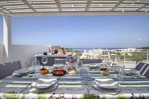 A dining table set outdoors with a view of the sea, a pergola overhead, and a terrace with white railings.