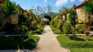 Un sentier bordé d’une végétation luxuriante et d’une architecture traditionnelle, qui mène à un bâtiment doté d’un toit en tuiles.