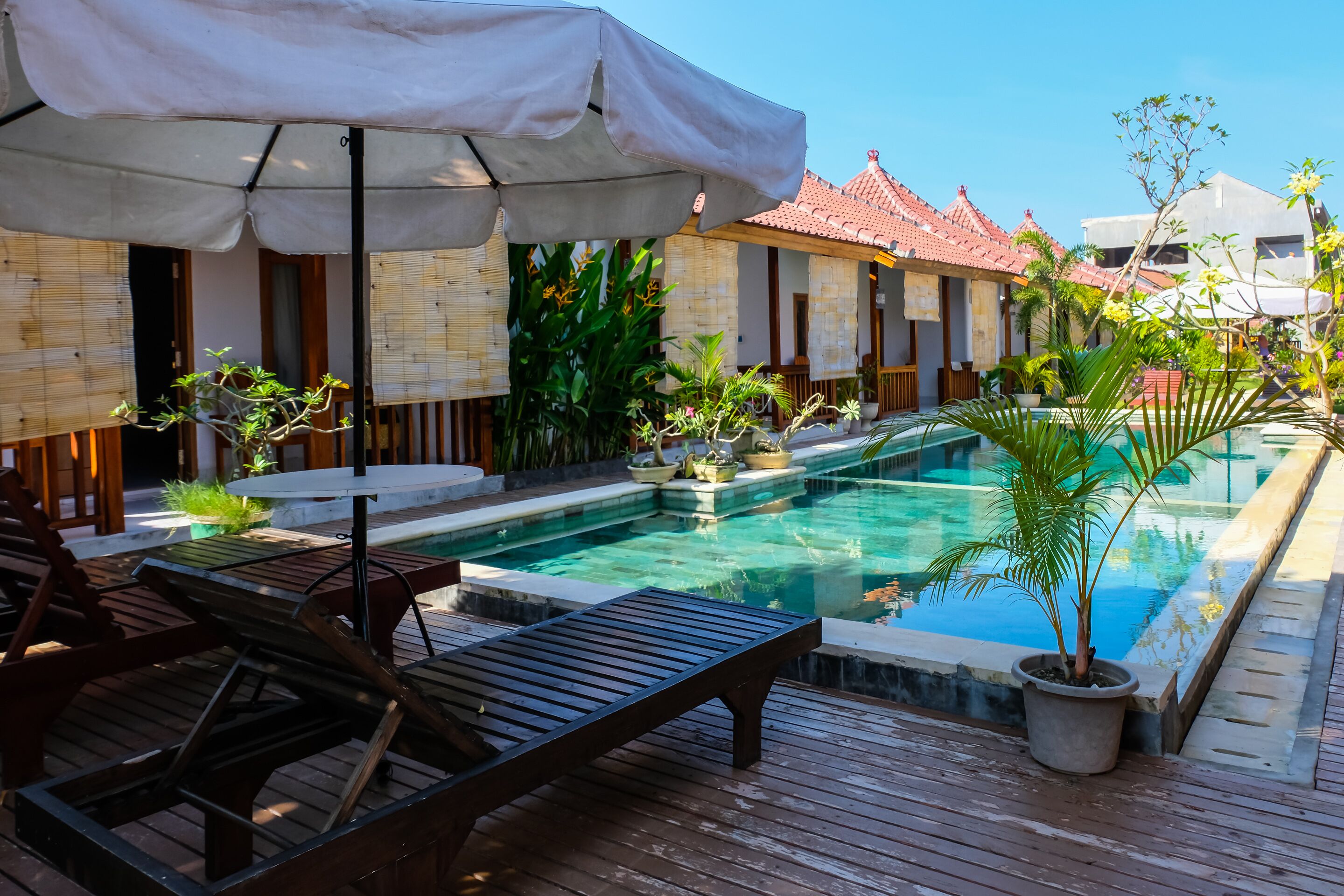 A pool area with lounge chairs and umbrellas, surrounded by potted plants and a traditional building with a tiled roof.