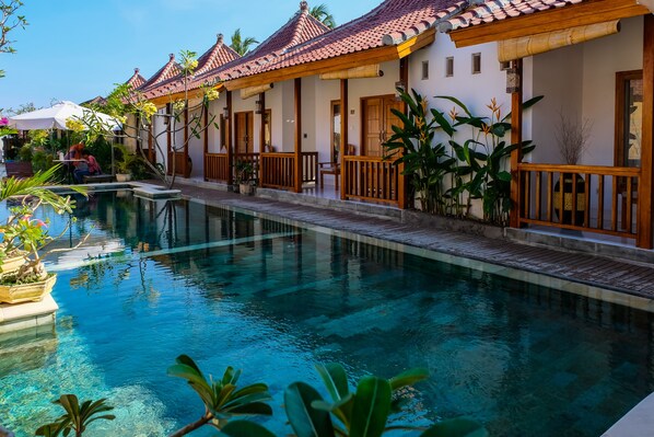 A poolside area with a clear pool, wooden deck, and traditional-style buildings with tiled roofs.