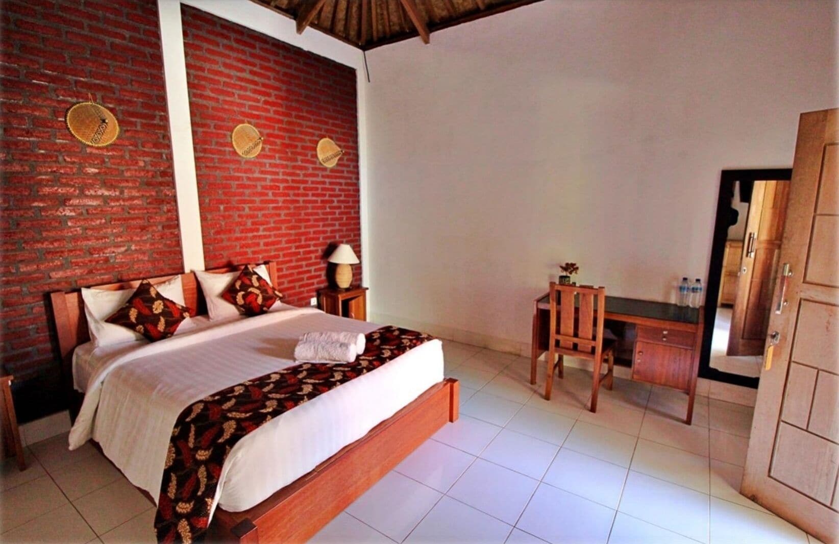A room with a brick wall, a bed with a patterned bedspread, a wooden desk, and a wardrobe.