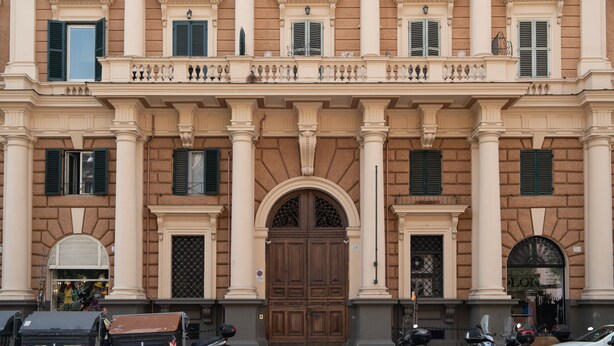 Ein Gebäude mit einem großen Eingangsbereich, zahlreichen Fenstern mit Fensterläden und einem Balkon mit Geländer.