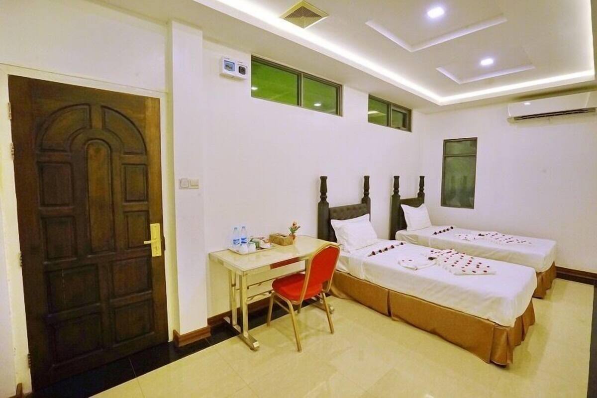Une chambre d’hôtel avec une porte en bois, un bureau avec une chaise, deux lits et une petite table sur laquelle se trouvent une bouteille et une boîte.