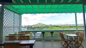 A covered outdoor seating area with tables and chairs, a ceiling fan, and a view of a cityscape with mountains in the distance. - Mandalay Hill View Inn (Mandalay)