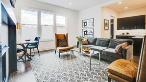A modern living room with a sofa, armchair, coffee table, and a dining area with chairs and a table. There are large windows providing natural light and a kitchen area with cabinets. - Close to Broadway · Rooftop Skyline · Walkable (Nashville)